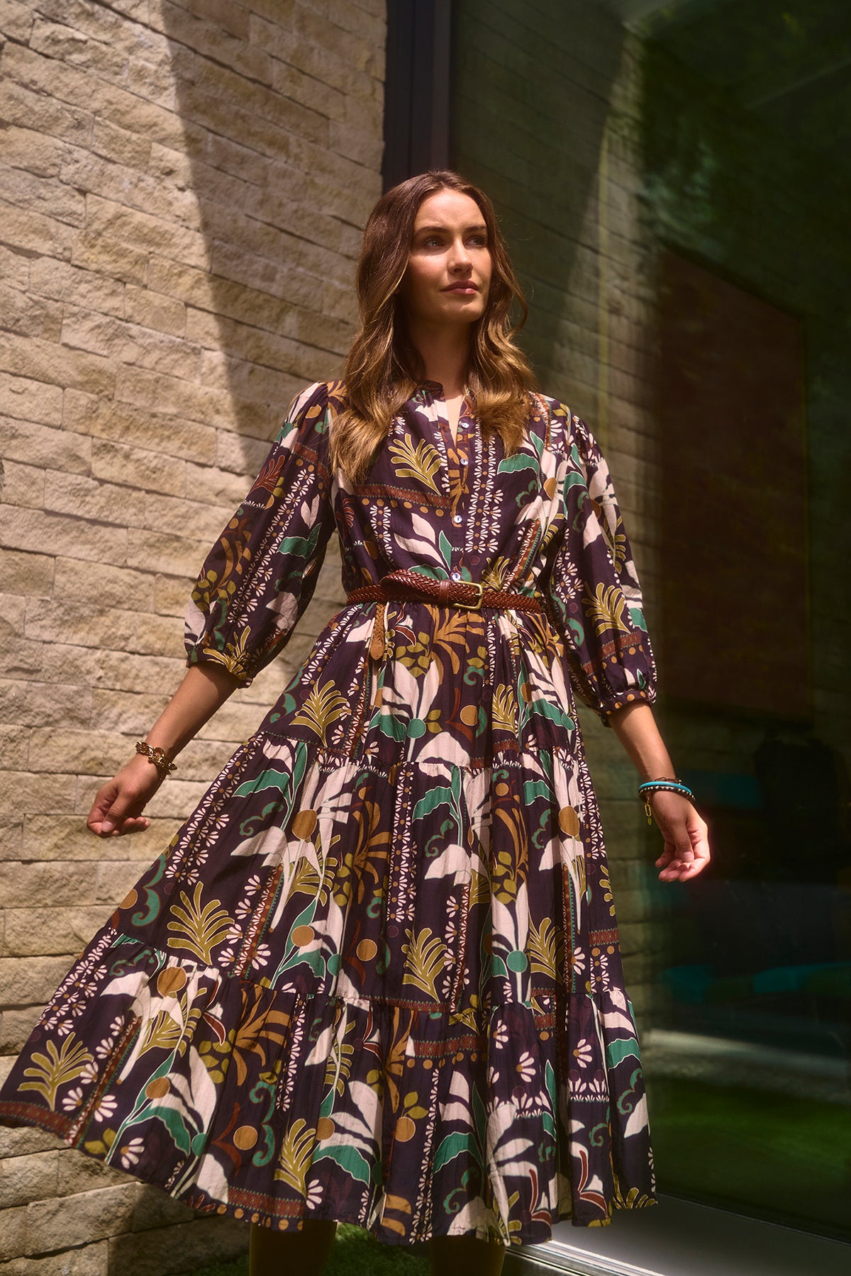   A woman stands against a brick wall wearing the JOHANNA PRINTED SILK COTTON VOILE DRESS from Velvet by Graham & Spencer, featuring a bold floral pattern, three-quarter sleeves, and a belt. She is looking to her right as sunlight casts shadows on the wall and her dress. 