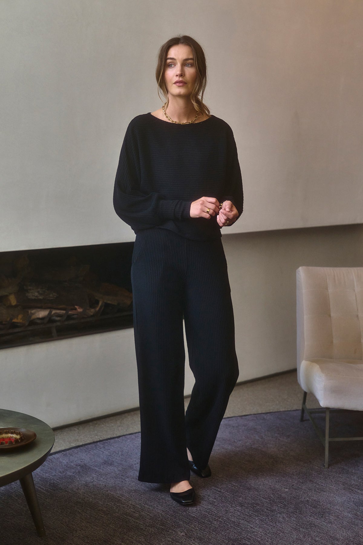   A woman dressed in a black outfit stands in a cozy room with a minimalist design. Beside her is a light-colored chair and a round table with a dish. The elegant touch of her attire is highlighted by her wide-leg trousers, which are the COLLEEN RIBBED PANT from Velvet by Graham & Spencer, made from a soft cotton blend. 