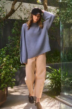 A person in a blue sweater and Velvet by Graham & Spencer's BRYLIE SANDED TWILL UTILITY PANT stands indoors by a large window and plants, wearing a black cap and loafers.