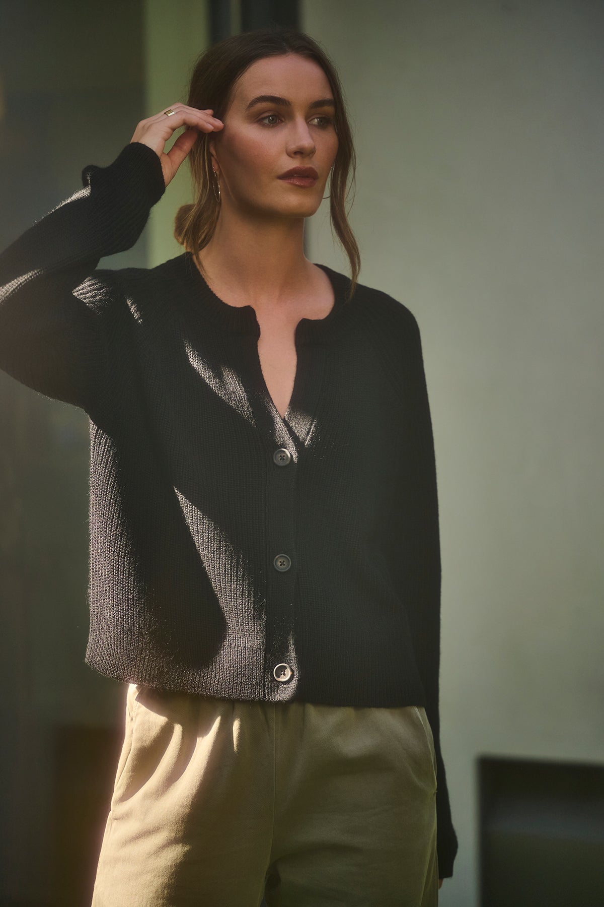   A person stands in a shadowed area, wearing the SHAYLA CARDIGAN by Velvet by Graham & Spencer—crafted from a cozy wool blend with raglan sleeves—and khaki pants, with one hand adjusting their hair. 