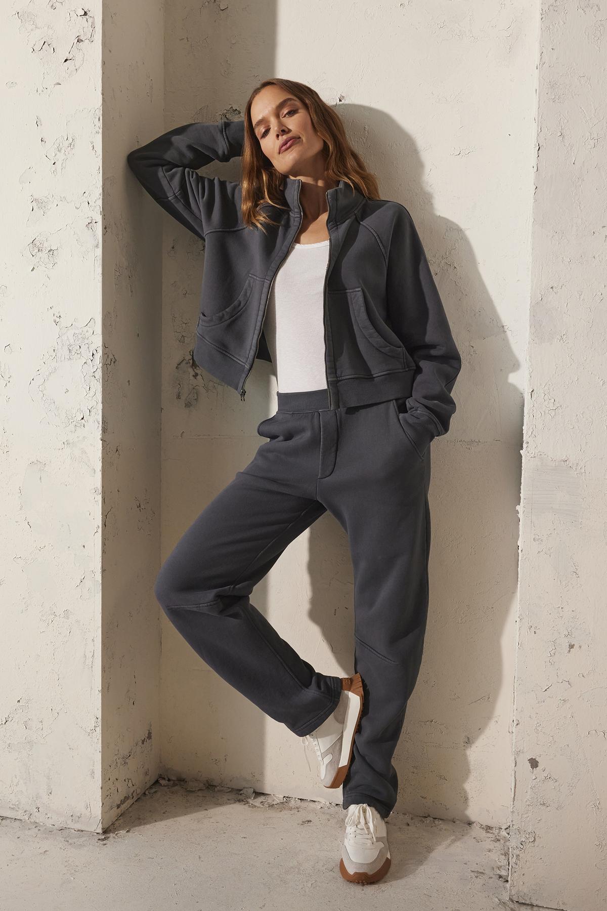   Wearing a cozy Velvet by Graham & Spencer CECILIA FULL-ZIP JACKET in charcoal, paired with white sneakers, a person casually leans against a textured wall with one hand up, exuding a relaxed vibe. 