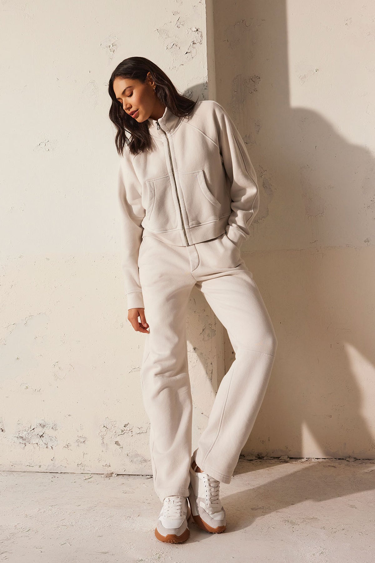   A person in a beige tracksuit and white sneakers, featuring the cozy CECILIA FULL-ZIP JACKET by Velvet by Graham & Spencer with raglan sleeves, leans against a textured cream wall. 