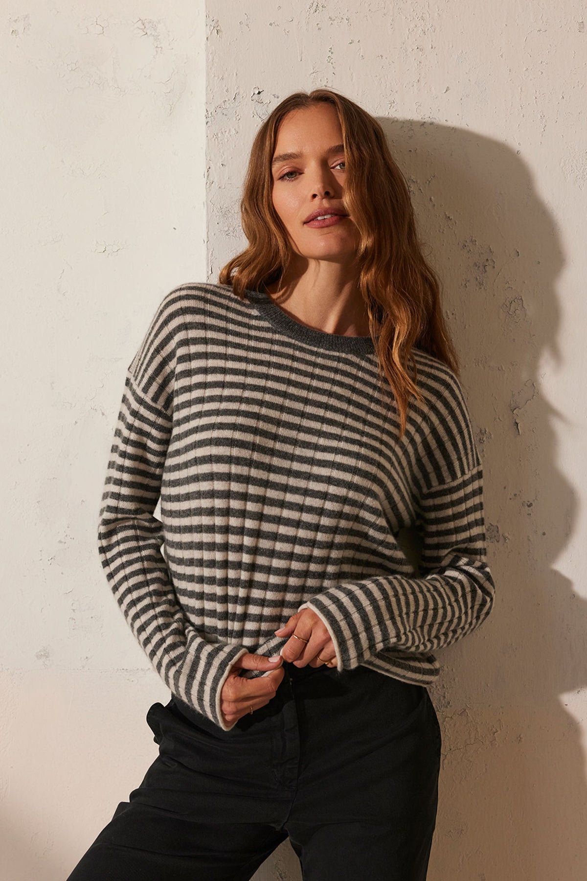 A person wearing the MONICA CASHMERE SWEATER by Velvet by Graham & Spencer and dark pants stands against a textured wall, exuding an air of luxurious comfort.-38301405184193