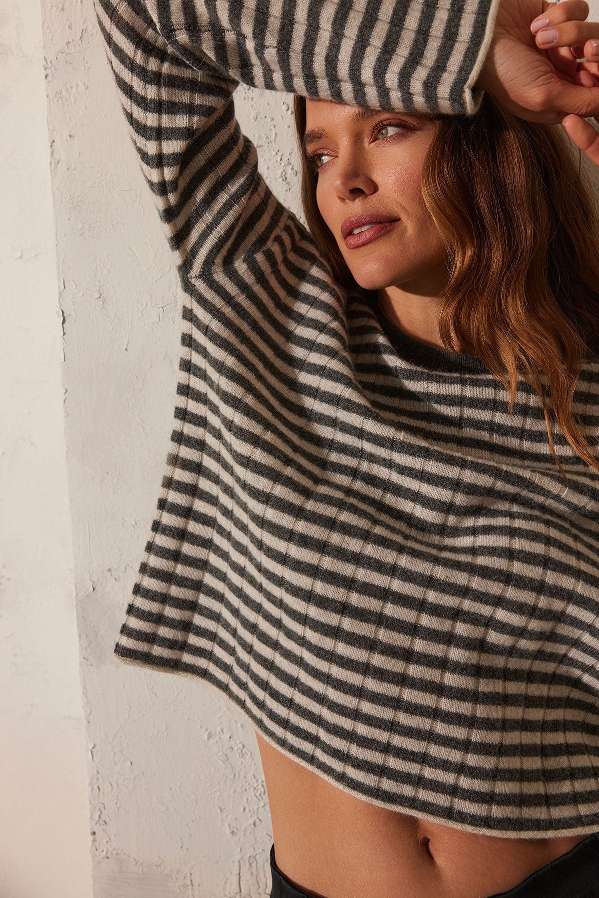   A person wearing the Velvet by Graham & Spencer MONICA CASHMERE SWEATER, with one arm raised, leans against a white textured wall, exuding both style and luxurious comfort. 