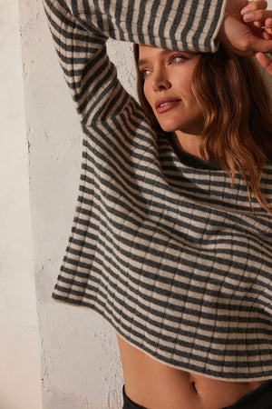 A person wearing the Velvet by Graham & Spencer MONICA CASHMERE SWEATER, with one arm raised, leans against a white textured wall, exuding both style and luxurious comfort.