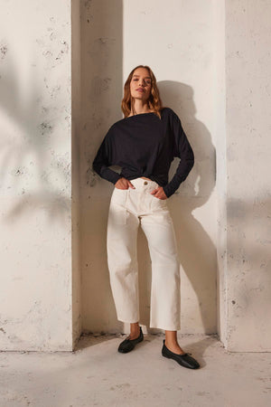 Against a textured white wall, a person wears a black long-sleeve shirt, the BRYLIE SANDED TWILL UTILITY PANT by Velvet by Graham & Spencer, and black shoes.