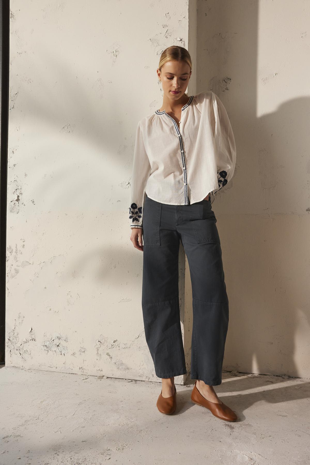   A person wearing the Velvet by Graham & Spencer BRYLIE SANDED TWILL UTILITY PANT and a white blouse stands against a textured wall, hands in the patch pockets. 