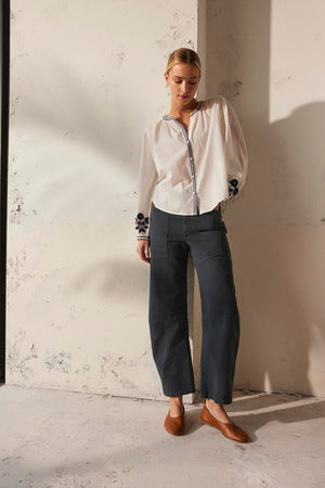 A person wearing the Velvet by Graham & Spencer BRYLIE SANDED TWILL UTILITY PANT and a white blouse stands against a textured wall, hands in the patch pockets.