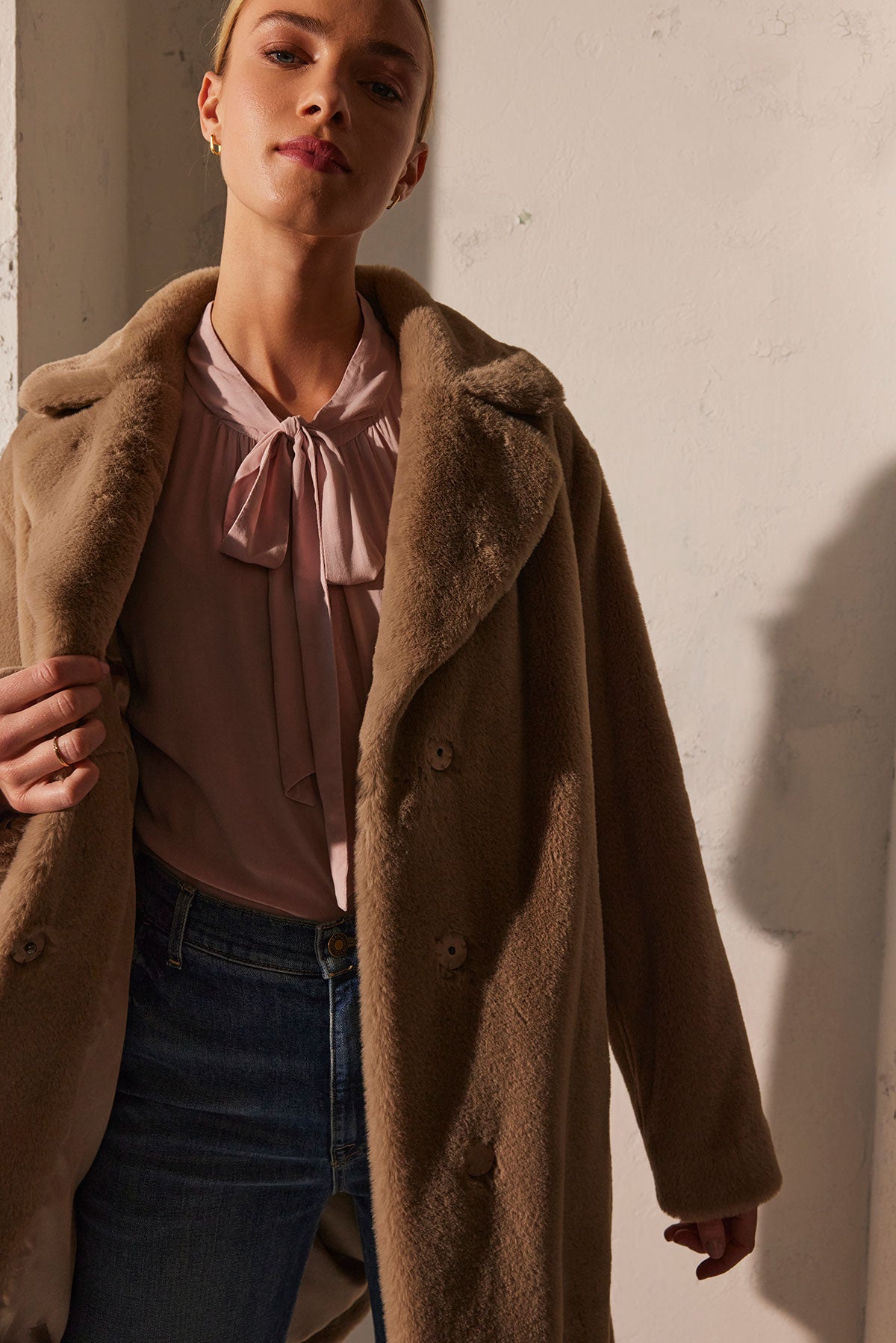   A woman wearing the GABRIELLE TOP by Velvet by Graham & Spencer, which features a pink blouse with a banded collar, stands against a light background. Her relaxed-fit beige coat drapes over her shoulders, and her jeans are visible as she gazes downward slightly. 