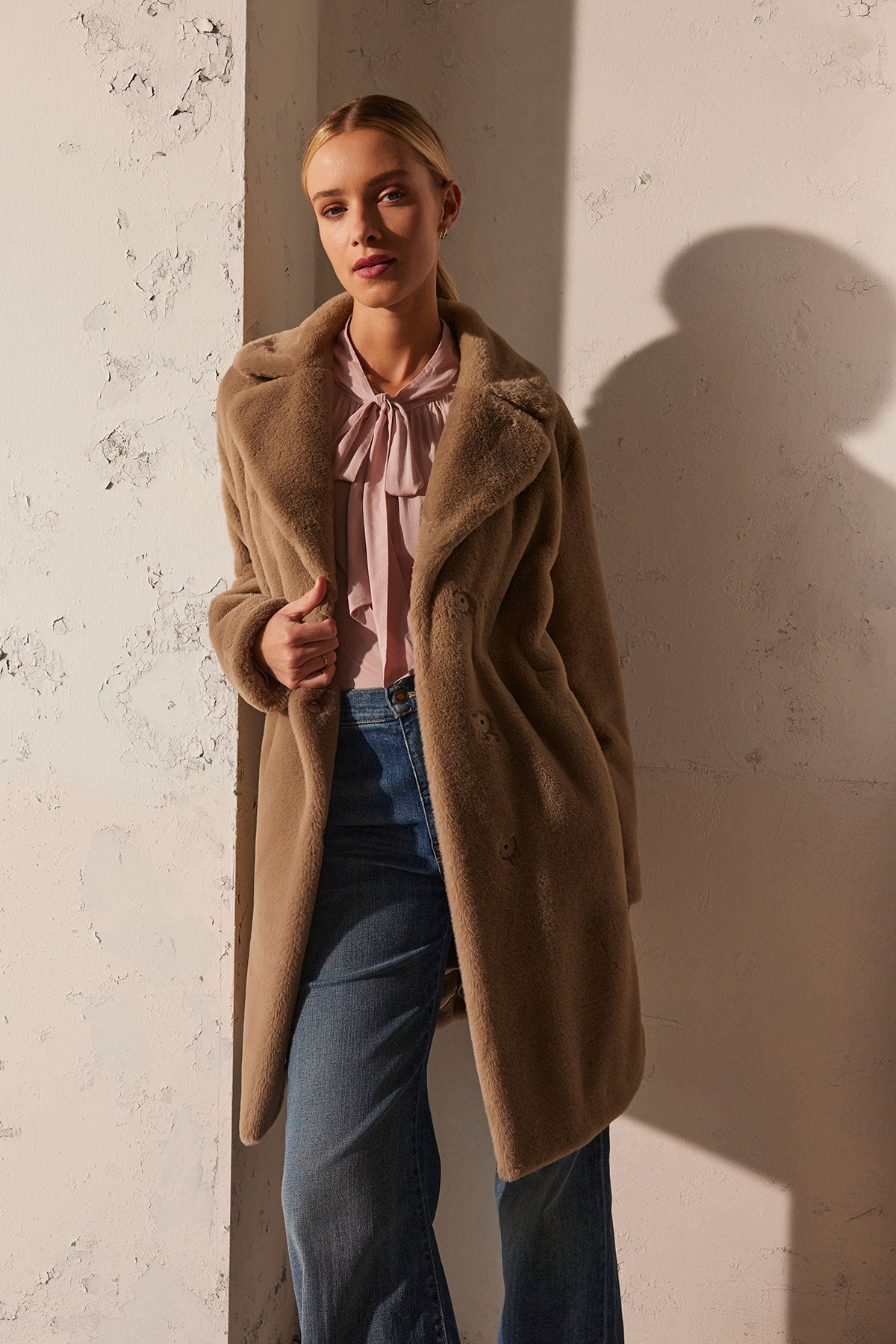   The individual is wearing the EVALYN LUX FAUX FUR COAT by Velvet by Graham & Spencer in tan, paired with a pink blouse and blue jeans, standing against a textured wall with a shadow cast on it—perfectly styled for a seamless day-to-night transition. 