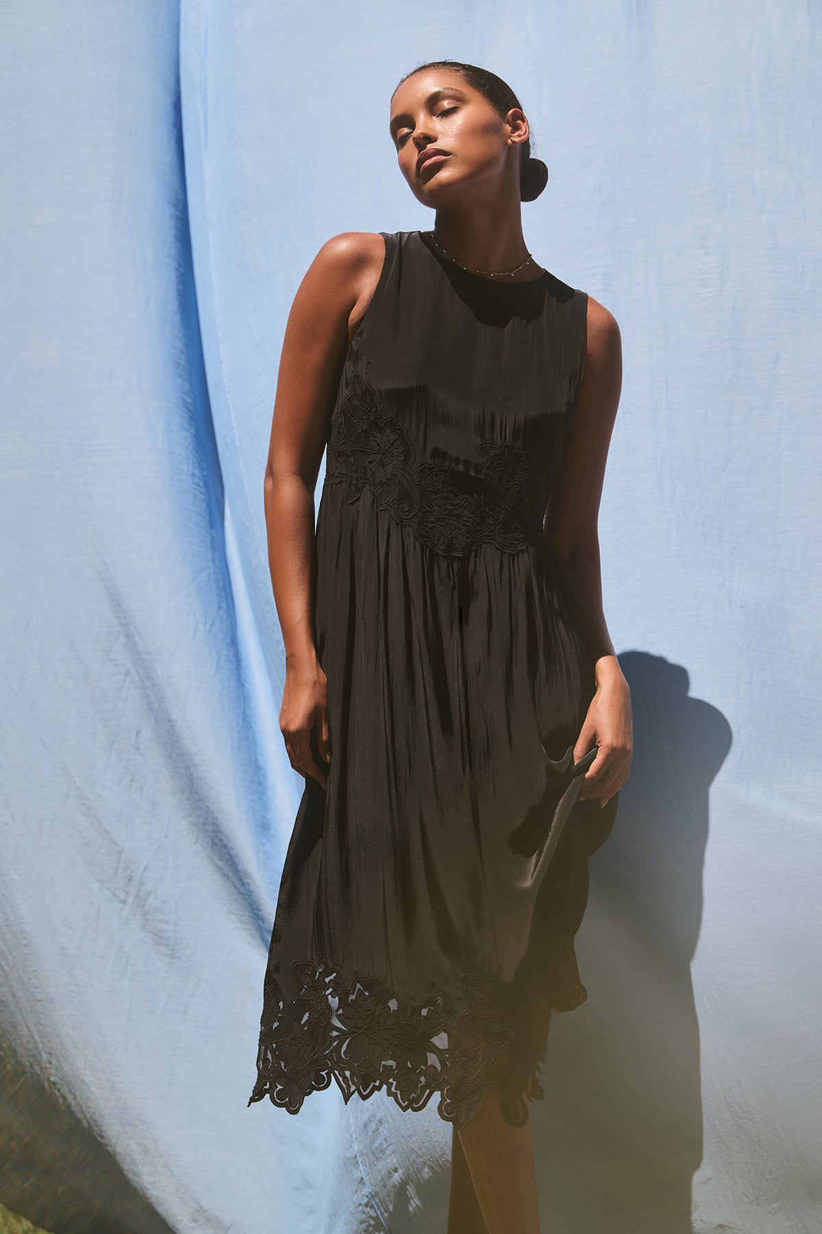   A person wearing the BIANCA DRESS by Velvet by Graham & Spencer, a sleeveless black dress featuring lace details, stands against a light blue fabric backdrop. 