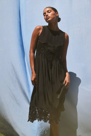 A person wearing the BIANCA DRESS by Velvet by Graham & Spencer, a sleeveless black dress featuring lace details, stands against a light blue fabric backdrop.