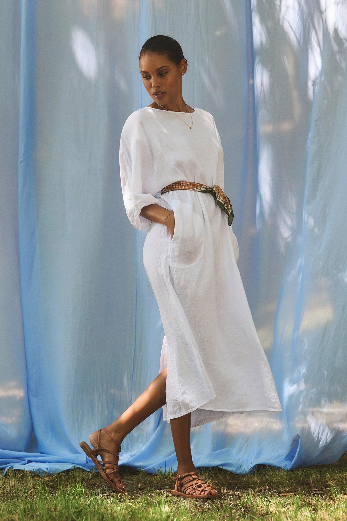   A person stands on grass in a Velvet by Graham & Spencer CARMEN LINEN DRESS, featuring dolman sleeves and sandals, against a flowing blue and white backdrop. 