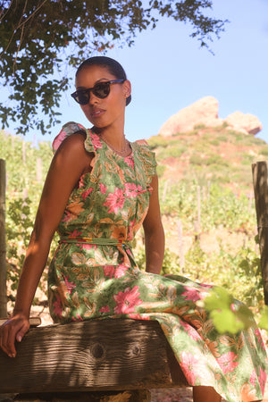 A woman wearing the ELARIA DRESS by Velvet by Graham & Spencer pairs sunglasses with a floral design as she sits on a wooden bench in a sunny outdoor setting with green foliage and distant rocks.