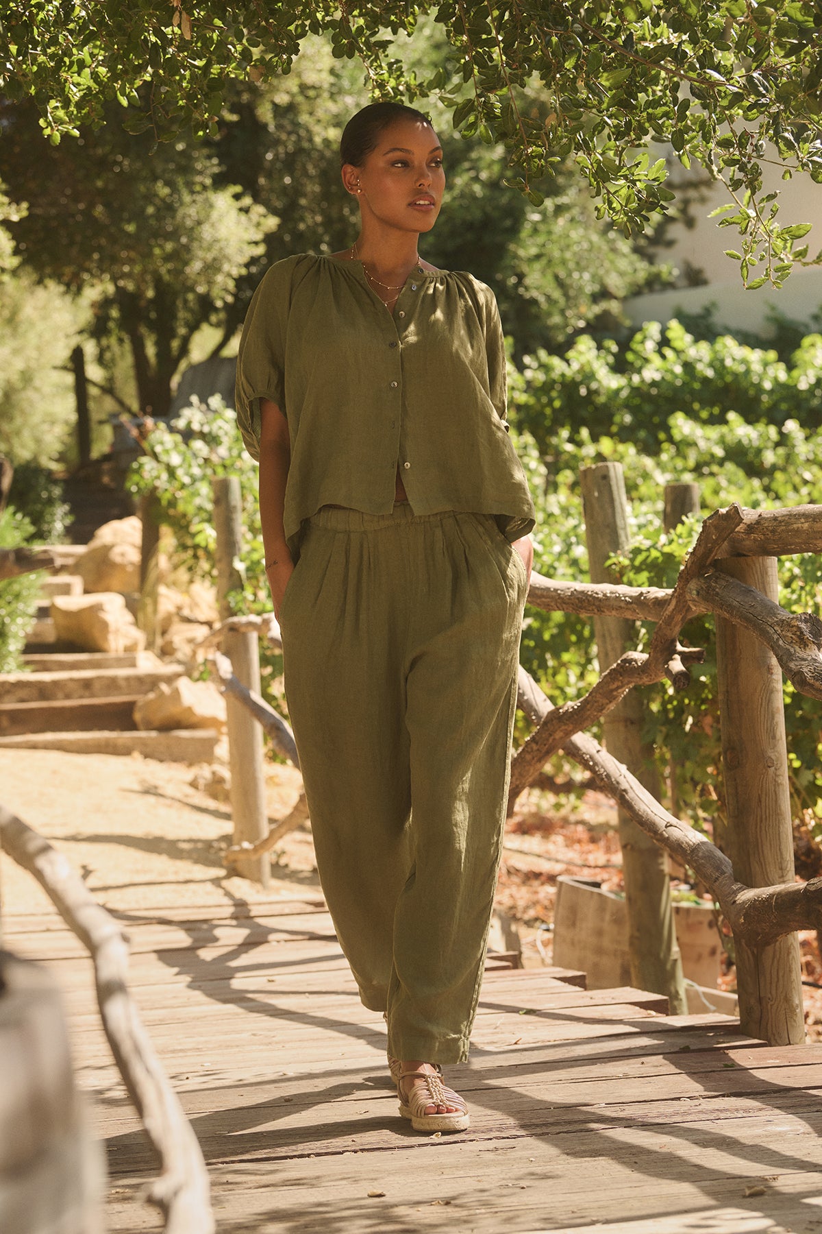 A person in the JAIMI LINEN TOP by Velvet by Graham & Spencer, featuring a button-front design and puff sleeves, stands on a wooden bridge enveloped in greenery with hands casually placed in pockets.-38662776324289