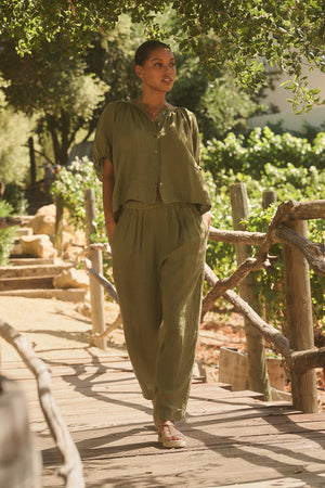 A person in the JAIMI LINEN TOP by Velvet by Graham & Spencer, featuring a button-front design and puff sleeves, stands on a wooden bridge enveloped in greenery with hands casually placed in pockets.