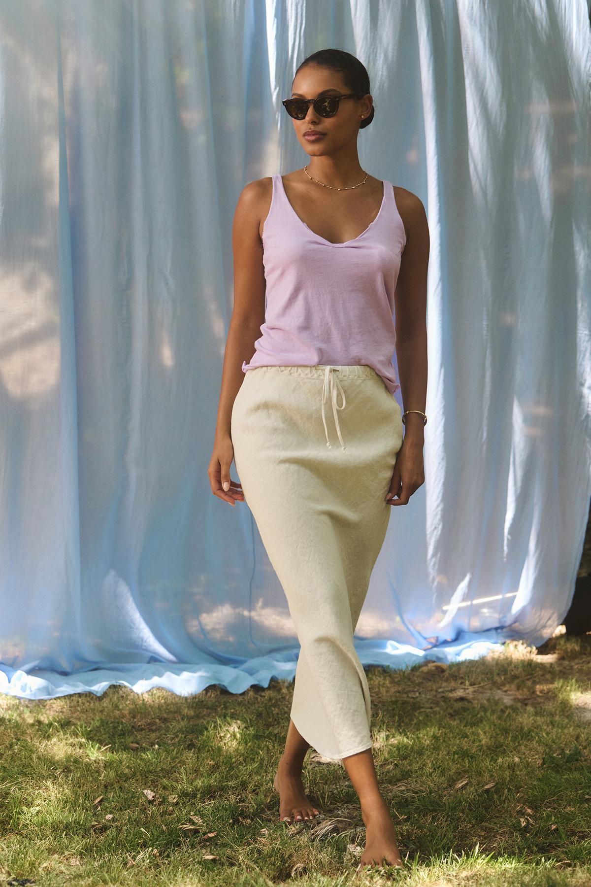   A woman wearing Velvet by Graham & Spencer's KIM Tank Top, sunglasses, and a cream skirt stands on grass in front of a light blue curtain backdrop. 