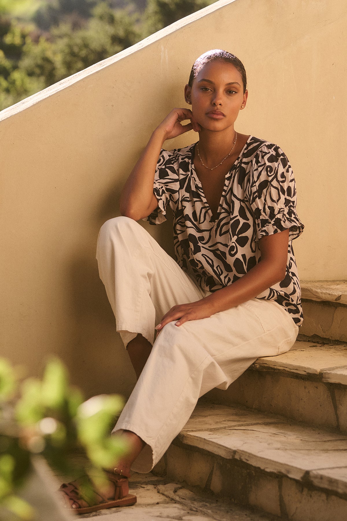   A person sits on outdoor steps wearing the Velvet by Graham & Spencer LESLIE TOP and light pants, resting their elbow on their knee, with greenery and a wall behind them. 