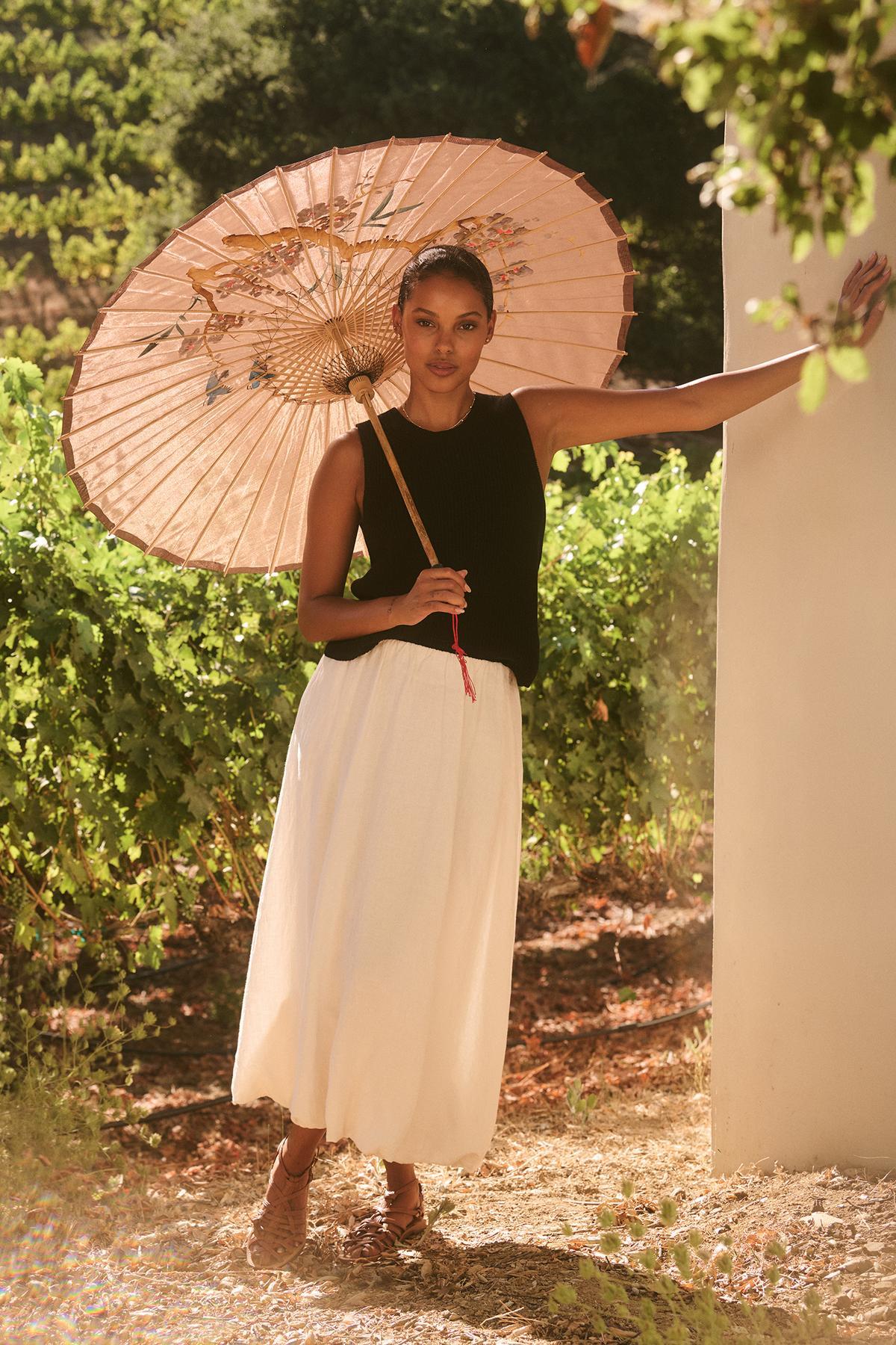   A person stands outdoors under a paper parasol, wearing the MUNA SWEATER VEST by Velvet by Graham & Spencer with a white skirt. They lean against a wall with greenery in the background. 