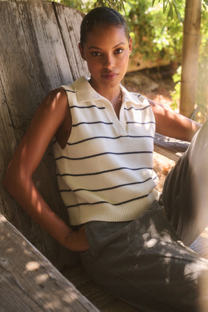 A person sits outdoors on a wooden bench, wearing the NAYLA COTTON CASHMERE SWEATER VEST by Velvet by Graham & Spencer, with lush greenery in the background.