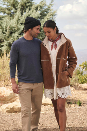 A man in a beanie and a Velvet by Graham & Spencer BECKER CREW sweater walks with a woman wearing a brown jacket and scarf. They are outdoors near a tree with hills in the background, enjoying the comfort of daily wear.