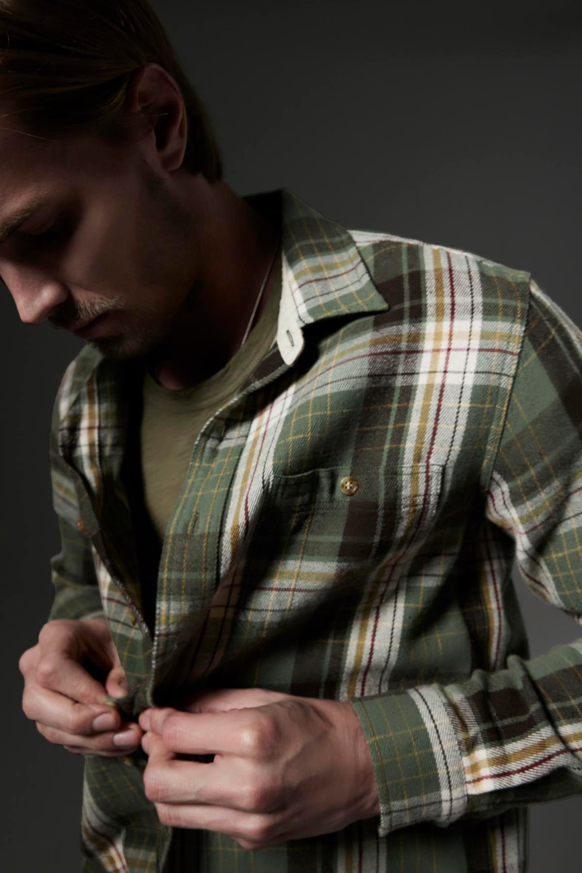   A person wearing a green plaid shirt is buttoning it up. With light hair and their gaze directed downwards, they stand against a dark gray background. This setting accentuates the quality of the Peruvian cotton fabric of the AMARO TEE by Velvet by Graham & Spencer, which features a classic crew neckline and provides the comfort of a lightweight tee. 