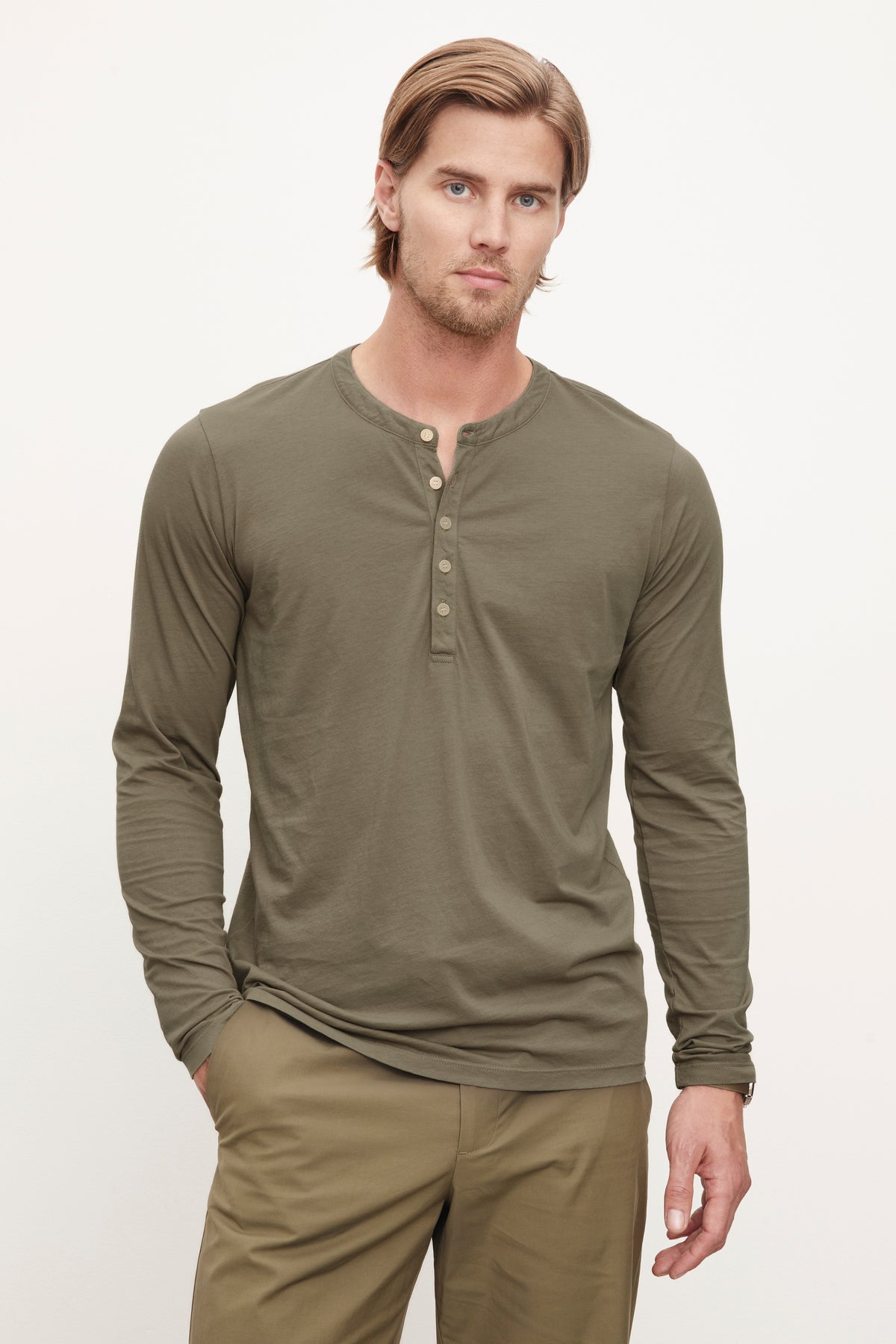   A man with blond hair and a beard, wearing a long-sleeve, olive green ALVARO HENLEY from Velvet by Graham & Spencer made from lightweight whisper knit fabric and khaki pants, stands with one hand in his pocket against a plain white background. 