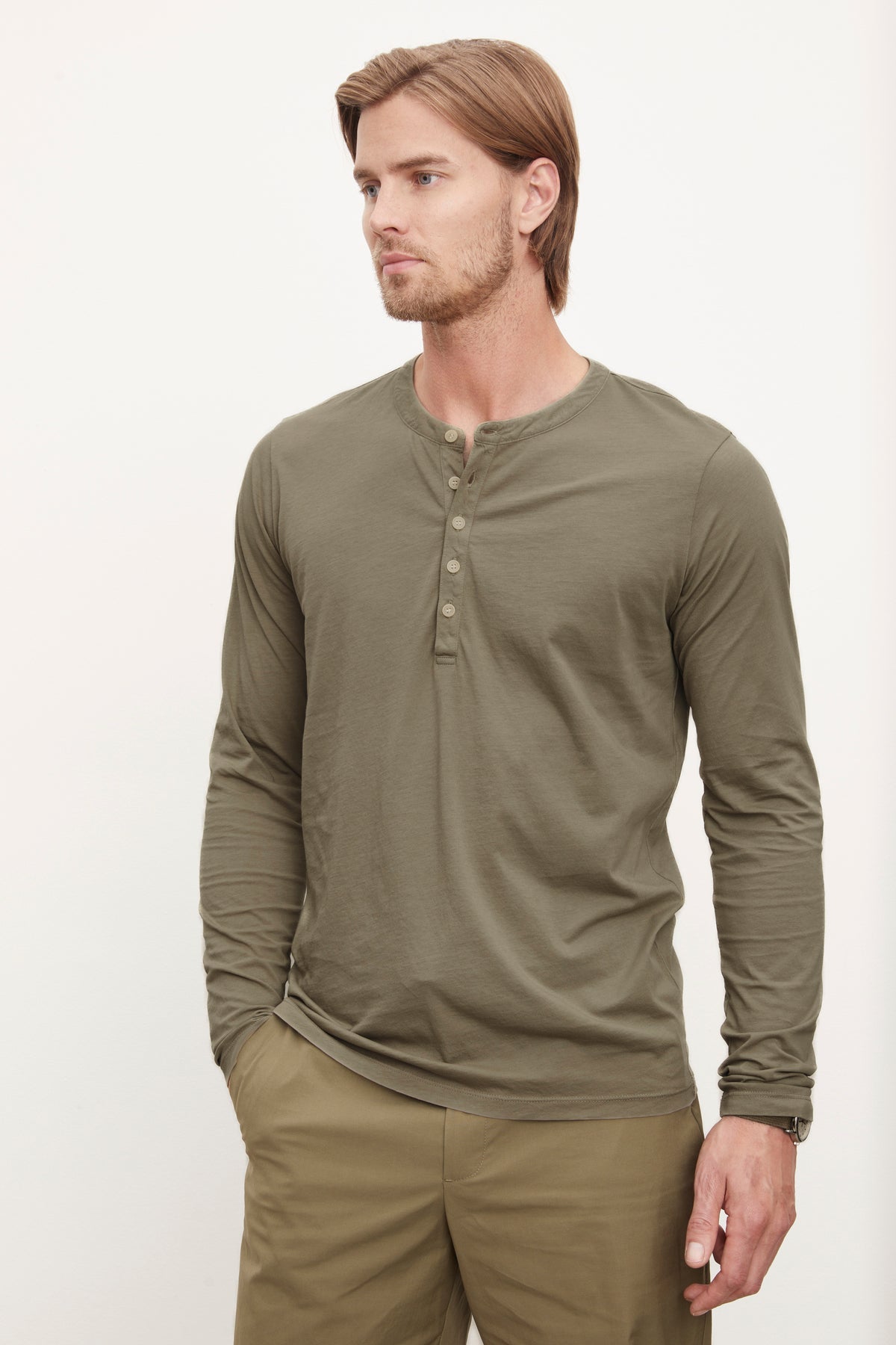   A man with medium-length hair stands wearing the Velvet by Graham & Spencer ALVARO HENLEY, a lightweight, long-sleeve olive green shirt with a buttoned henley neckline, and khaki pants. 
