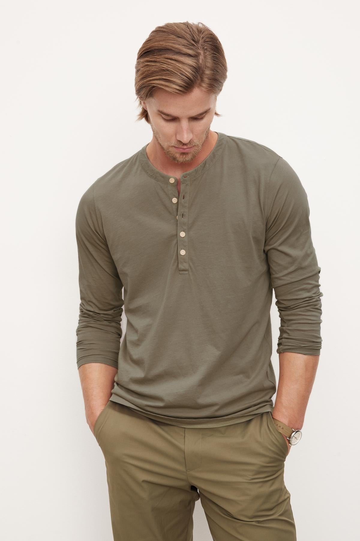   A man with light brown hair stands with hands in his pockets, looking down, wearing an olive green long-sleeve ALVARO HENLEY by Velvet by Graham & Spencer crafted from whisper knit material and matching pants against a plain white background. 