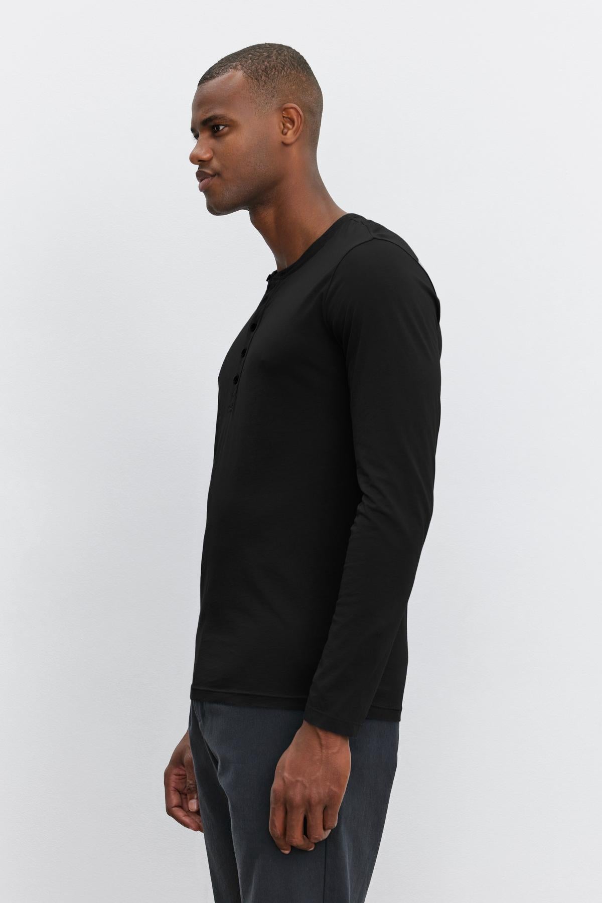   A man wearing a black lightweight ALVARO HENLEY by Velvet by Graham & Spencer and dark trousers stands against a plain white background, looking to the left. 