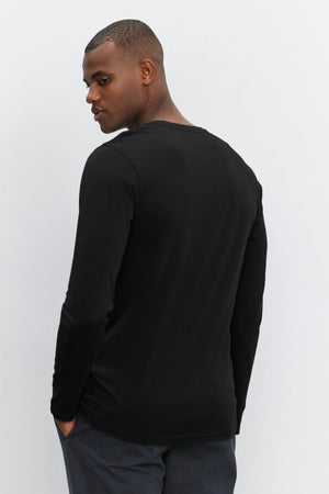 A person with short hair is standing and looking over their shoulder. They are wearing a long-sleeve black ALVARO HENLEY by Velvet by Graham & Spencer and dark pants against a plain white background.