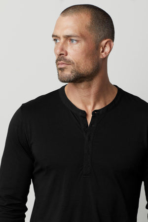 A man with a shaved head and beard is wearing a black ALVARO HENLEY by Velvet by Graham & Spencer, looking off to the side against a plain background.