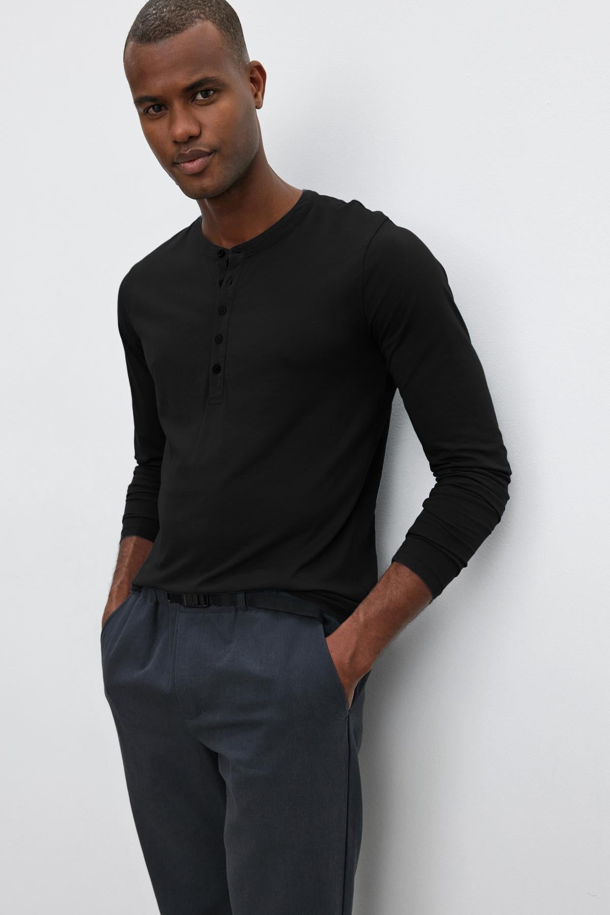   A person stands against a plain background, wearing a black long-sleeve ALVARO HENLEY by Velvet by Graham & Spencer and dark pants, with hands in pockets. 
