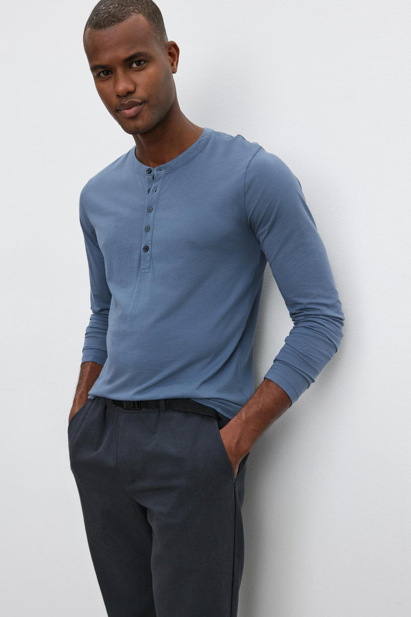 A man with short hair stands casually with hands in pockets, wearing the ALVARO HENLEY by Velvet by Graham & Spencer— a lightweight blue henley shirt that's perfect for layering. He pairs it with dark pants, all set against a plain white background.