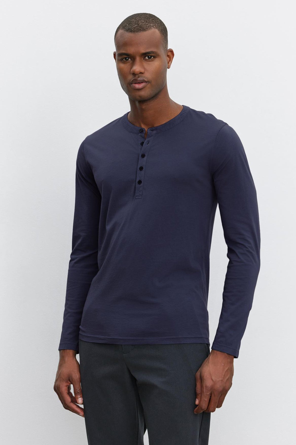 A man stands against a plain background, wearing a long-sleeve, dark blue ALVARO HENLEY by Velvet by Graham & Spencer and dark grey pants. The lightweight whisper knit fabric ensures comfort and style.-36273890525377