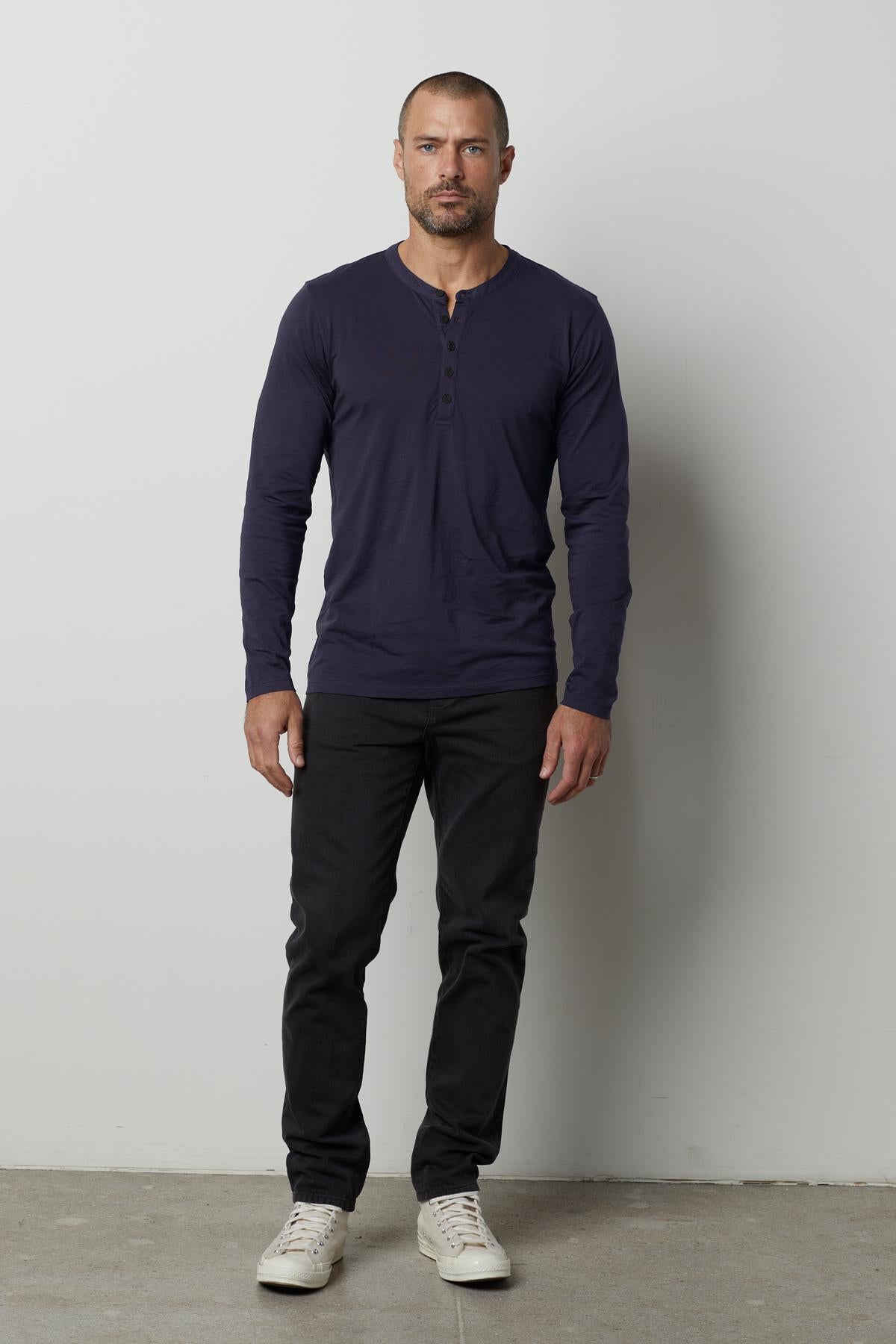 A man with a shaved head, wearing a long-sleeve lightweight navy ALVARO HENLEY by Velvet by Graham & Spencer, black jeans, and white sneakers, stands against a plain white background.-35607611113665
