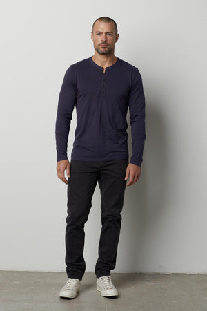 A man with a shaved head, wearing a long-sleeve lightweight navy ALVARO HENLEY by Velvet by Graham & Spencer, black jeans, and white sneakers, stands against a plain white background.