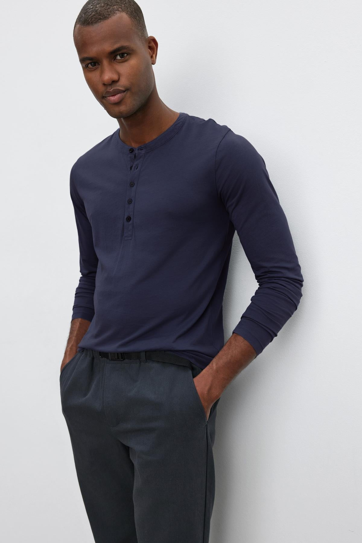   A man with short hair wears a navy long-sleeve ALVARO HENLEY by Velvet by Graham & Spencer and dark trousers, standing with his hands in his pockets against a plain white background. 