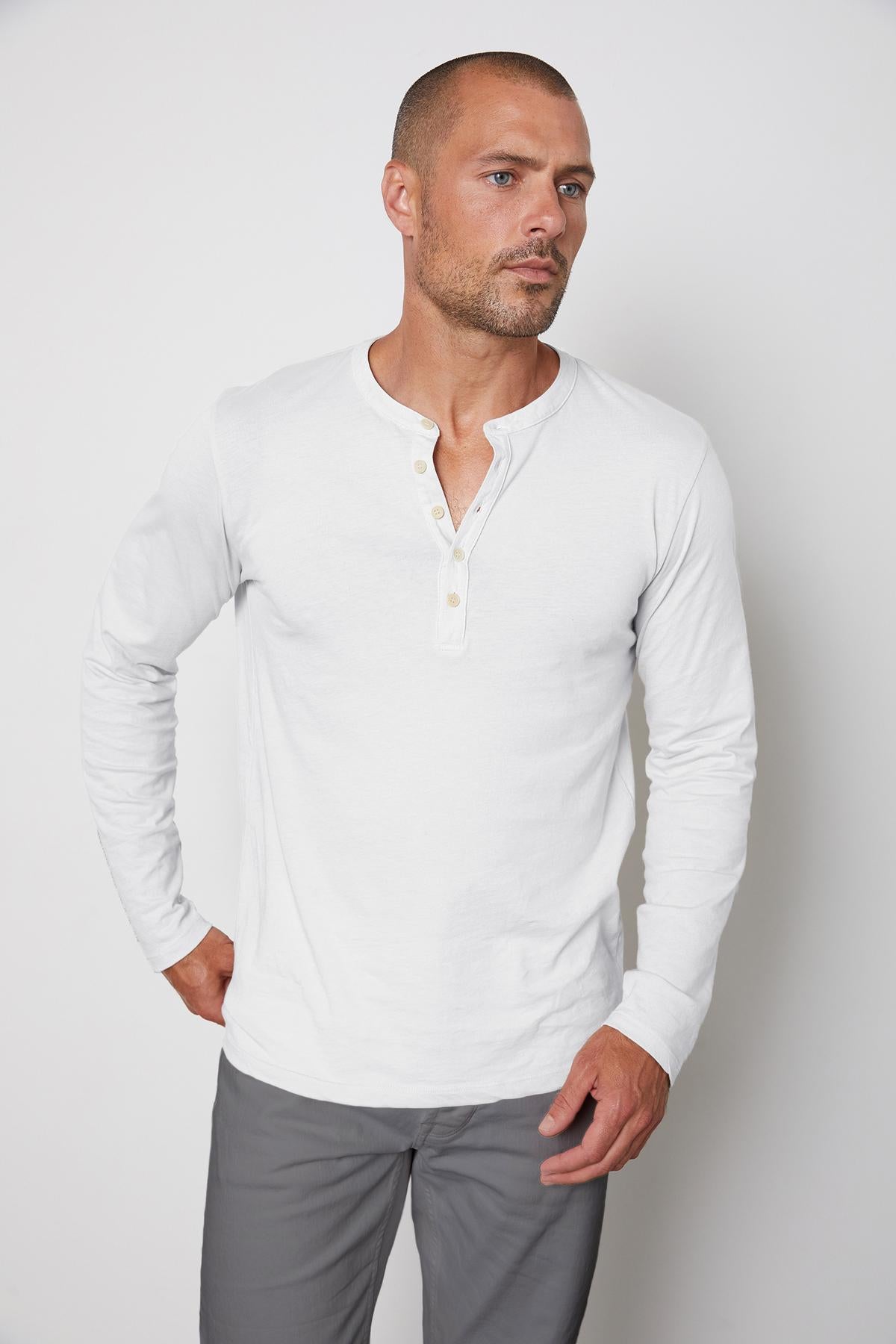 A man with a shaved head and short beard is wearing a white, long-sleeve ALVARO HENLEY shirt by Velvet by Graham & Spencer made from whisper knit fabric and gray pants. He stands with one hand on his hip, looking slightly to the side against a plain background.-36273890296001