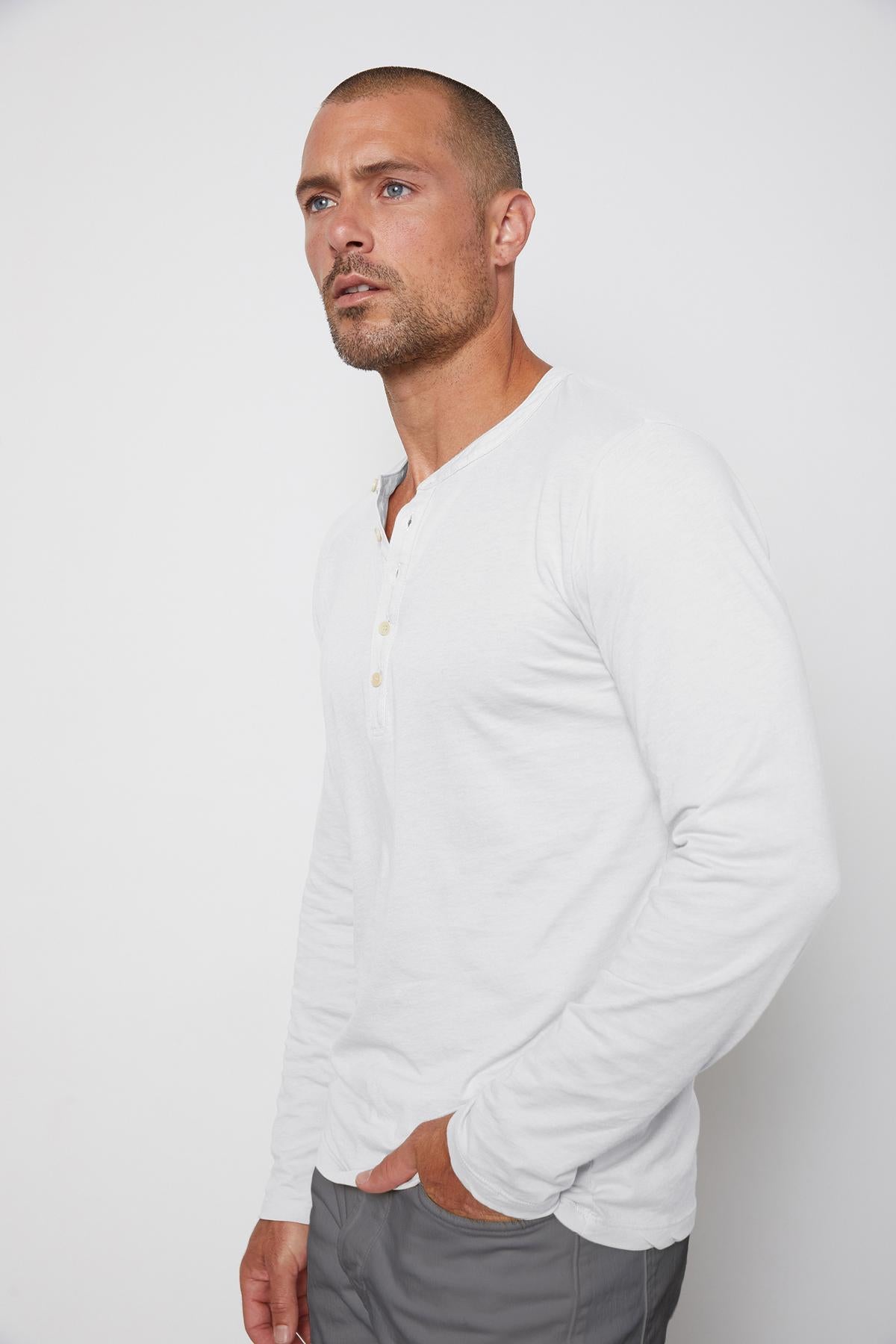 A man with a shaved head and a short beard wears a white long-sleeve ALVARO HENLEY by Velvet by Graham & Spencer and gray pants, standing against a plain white background.-36273890328769