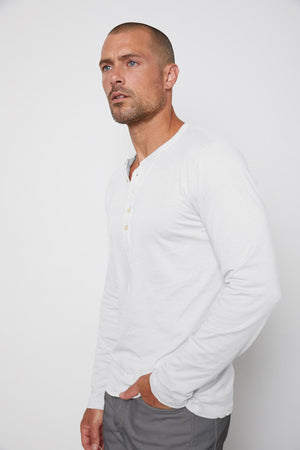 A man with a shaved head and a short beard wears a white long-sleeve ALVARO HENLEY by Velvet by Graham & Spencer and gray pants, standing against a plain white background.