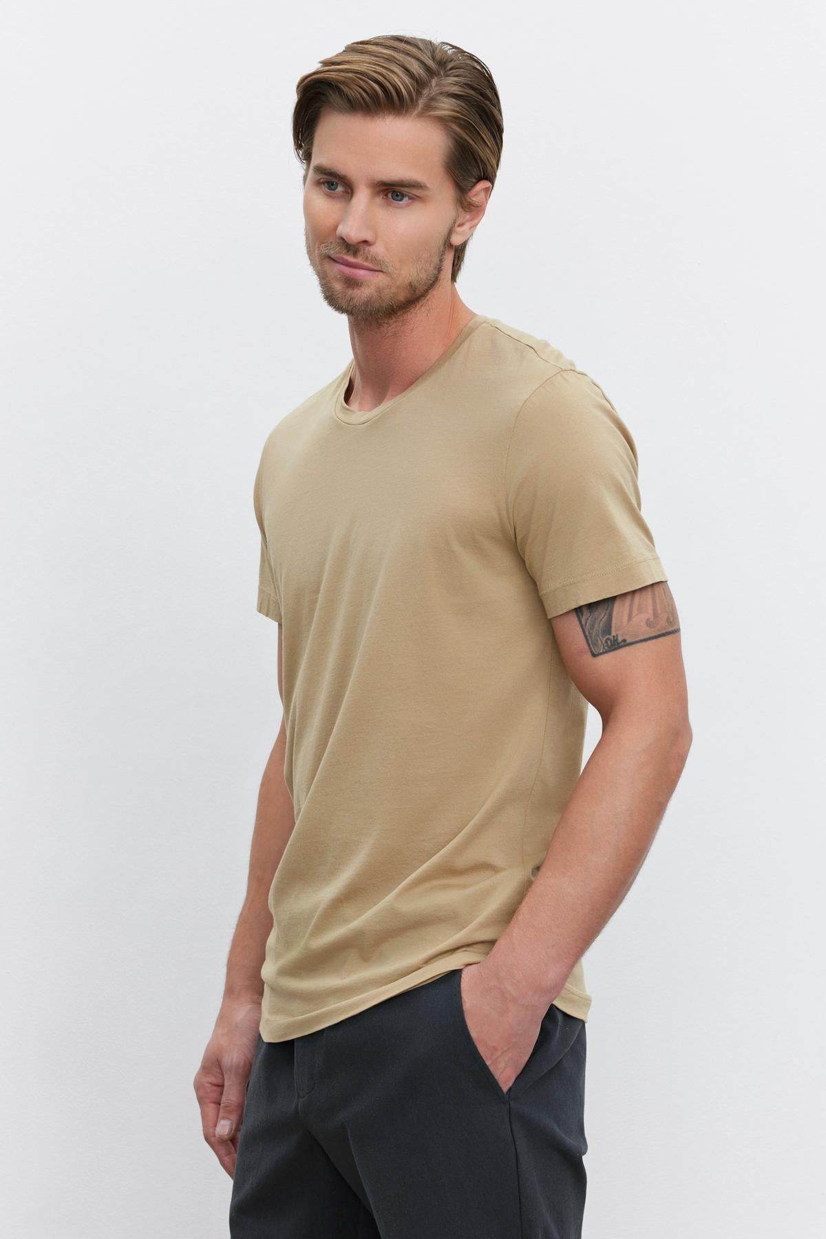 A man with short, styled hair and a tattoo on his left arm wearing a HOWARD TEE from Velvet by Graham & Spencer made of lightweight cotton knit and dark pants, standing with his hands in his pockets.-37386158801089