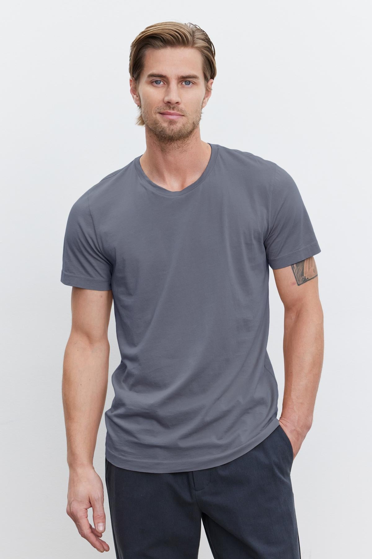   A person wearing a gray HOWARD TEE made of lightweight cotton knit from Velvet by Graham & Spencer and dark pants stands against a plain white background. One arm is resting by their side while the other is partially in their pocket, creating a relaxed yet perfect fit with a hint of vintage feel. 