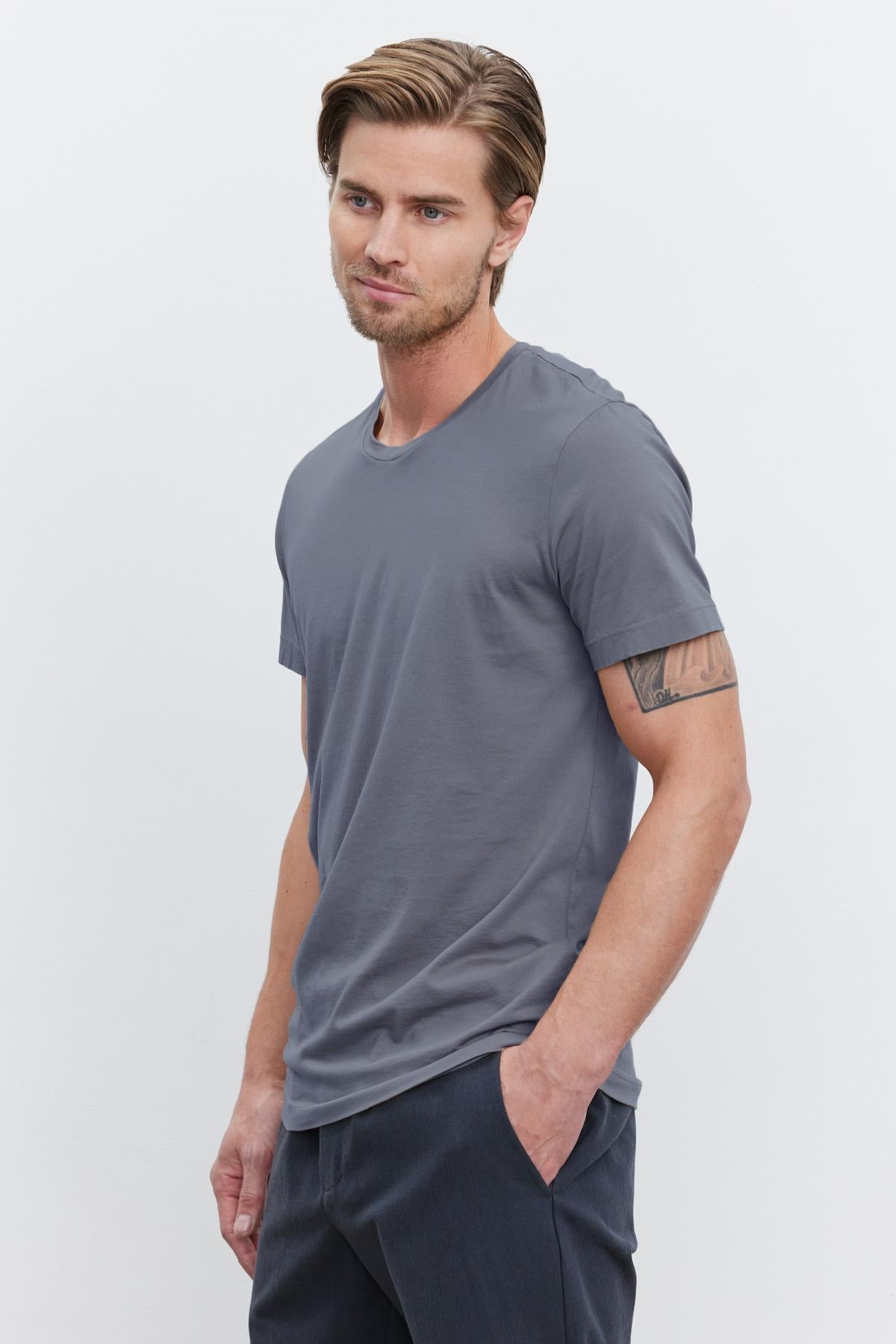   A man with light brown hair and a beard wearing a gray HOWARD TEE made of lightweight cotton knit by Velvet by Graham & Spencer and dark pants stands against a plain white background, exuding a vintage feel with one hand in his pocket. 