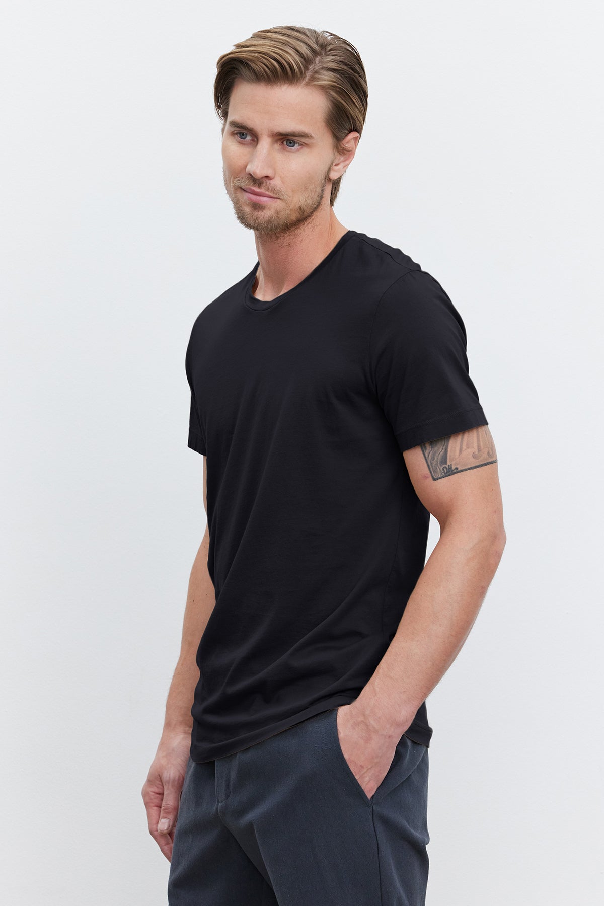   A man with short hair, a beard, and a tattoo on his right arm, wearing a black HOWARD TEE by Velvet by Graham & Spencer made from lightweight cotton knit and gray pants that give off a vintage feel, with his left hand in his pocket, stands against a plain white background. 