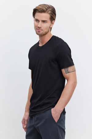 A man with short hair, a beard, and a tattoo on his right arm, wearing a black HOWARD TEE by Velvet by Graham & Spencer made from lightweight cotton knit and gray pants that give off a vintage feel, with his left hand in his pocket, stands against a plain white background.