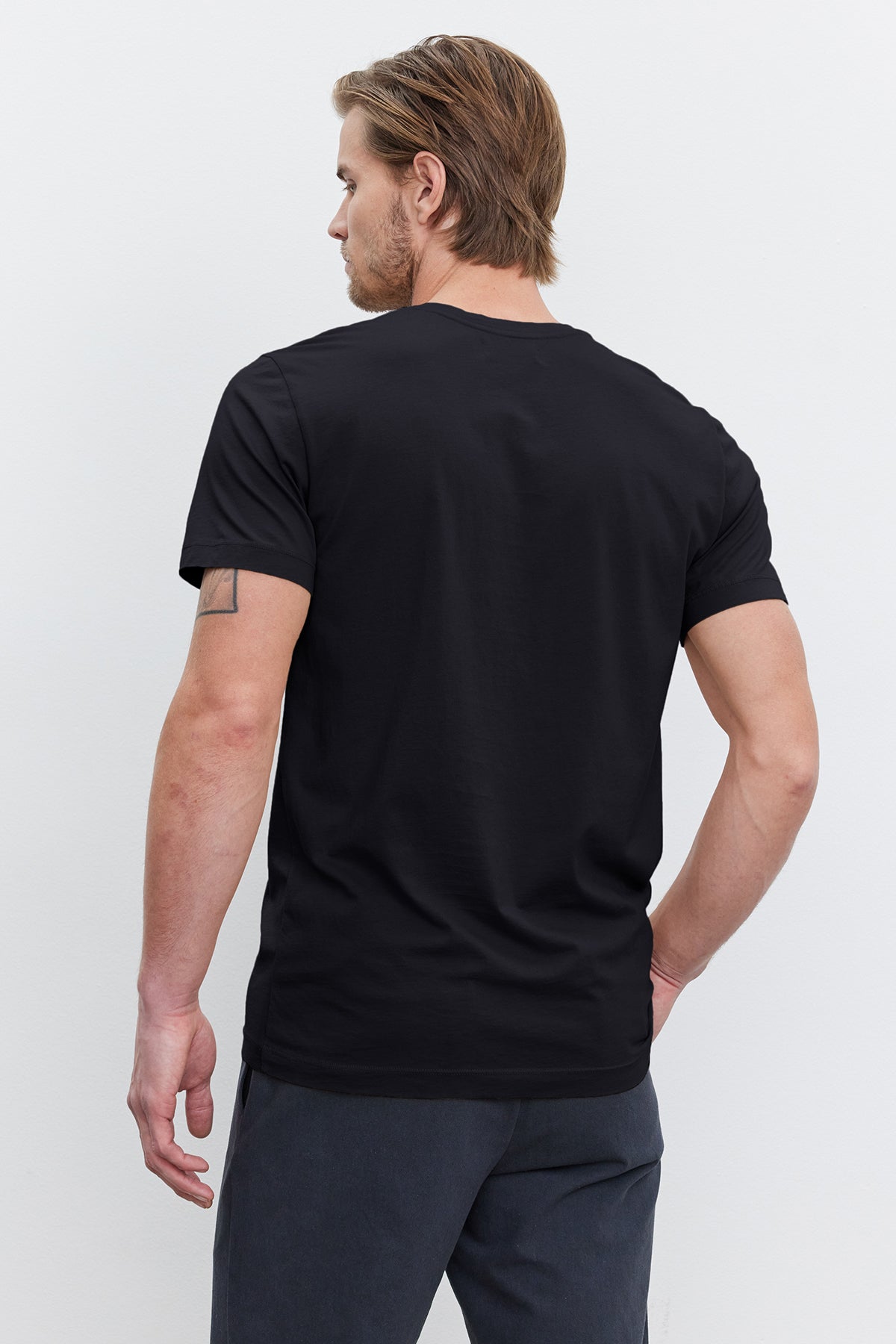   A man with short brown hair is seen from the back, wearing a black HOWARD TEE by Velvet by Graham & Spencer and dark pants. His lightweight cotton knit shirt offers a perfect fit, and he has a tattoo on his left arm. 