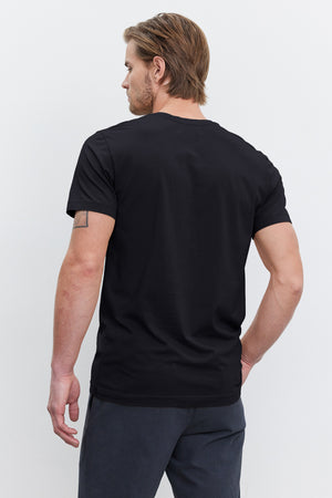 A man with short brown hair is seen from the back, wearing a black HOWARD TEE by Velvet by Graham & Spencer and dark pants. His lightweight cotton knit shirt offers a perfect fit, and he has a tattoo on his left arm.