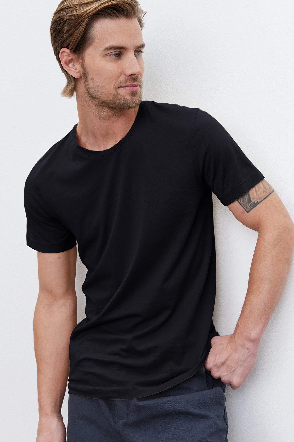   A man with light brown hair and a short beard, wearing a plain black HOWARD TEE made of lightweight cotton knit by Velvet by Graham & Spencer and dark pants, poses against a white background. He has a tattoo on his left forearm and is looking to his left, giving the image a vintage feel. 