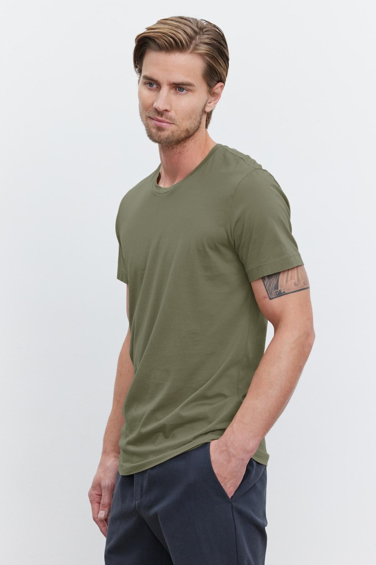   A young man with light brown hair wearing an olive green HOWARD TEE from Velvet by Graham & Spencer made from lightweight cotton knit and dark pants stands with his hands in his pockets against a plain white background, giving off a perfect fit. 